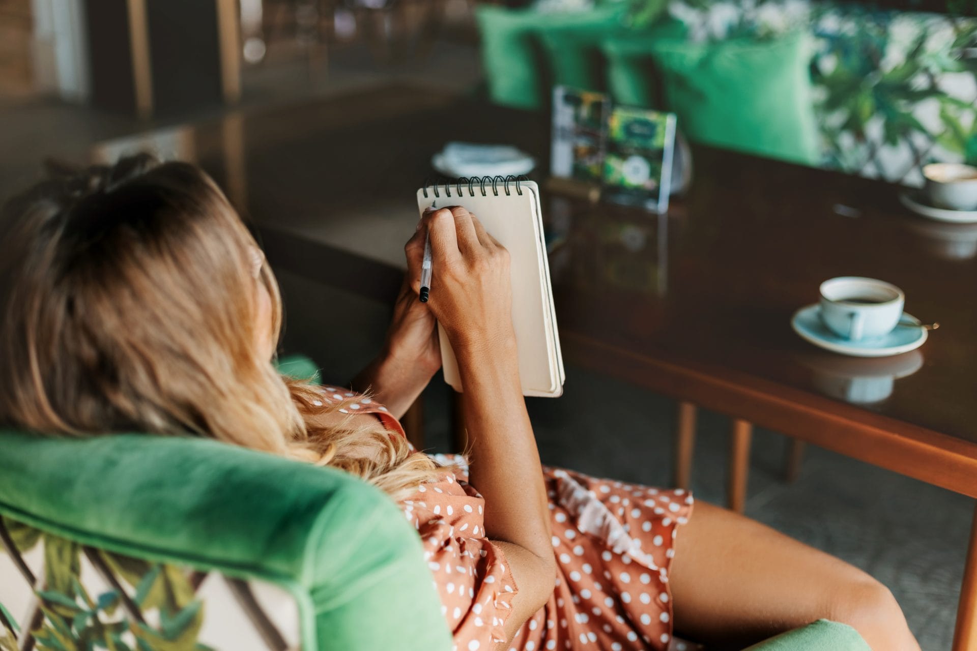 Woman sitting in a green armchair writing in a spiral notebook, with a cup of coffee on the table nearby, reflecting on relapse triggers in recovery
